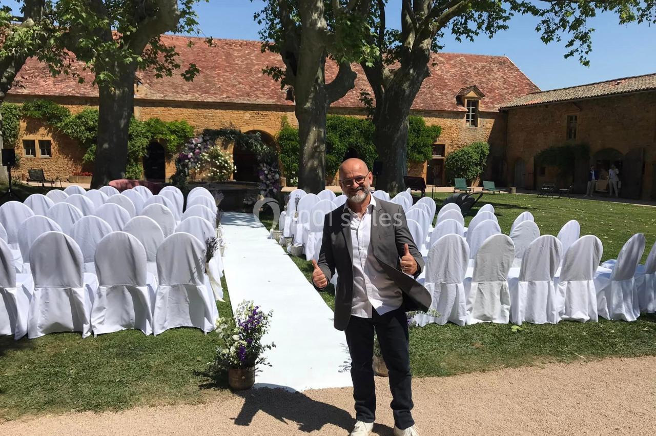 Homme souriant debout devant une allée centrale bordée de chaises blanches, dans un jardin avec bâtiments en pierre.