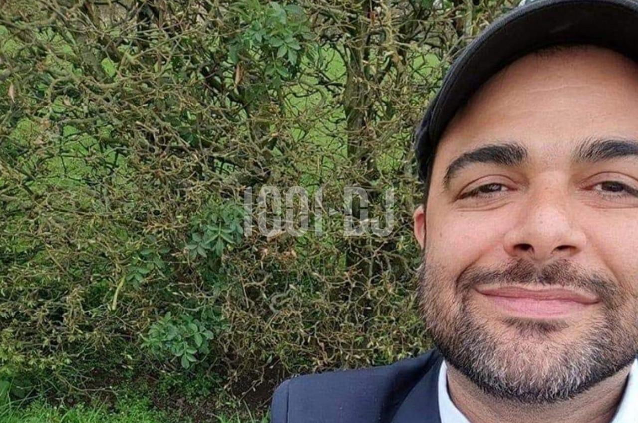 Un homme souriant portant une casquette devant un buisson dense dans un environnement extérieur.