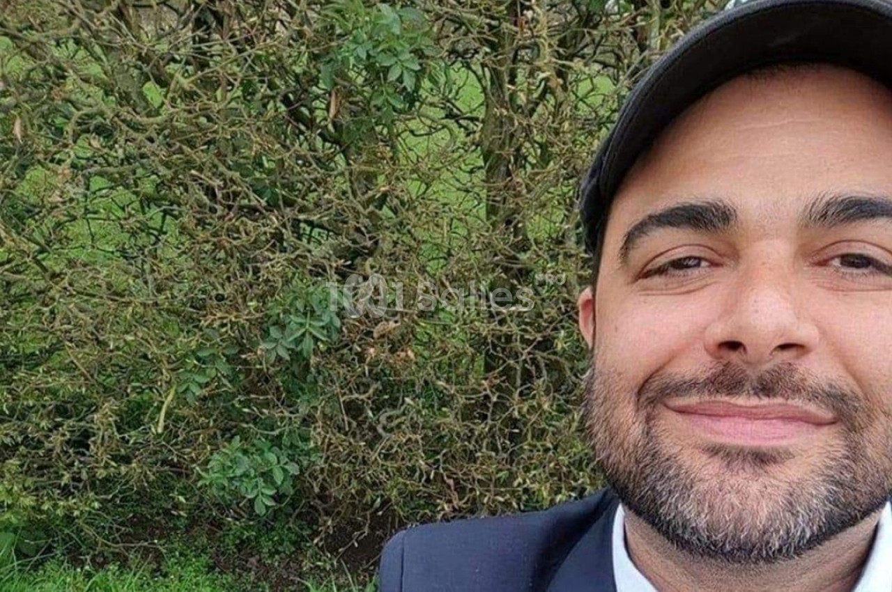 Un homme souriant portant une casquette devant un buisson dense dans un environnement extérieur.