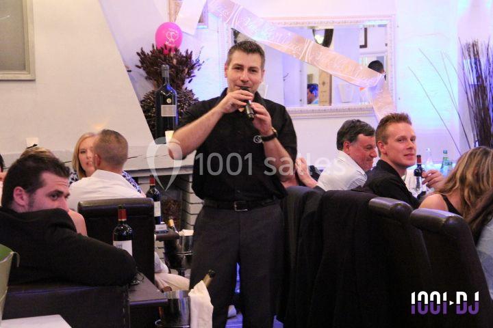 Un homme en chemise noire parle au micro dans un restaurant, entouré de convives assis à des tables.