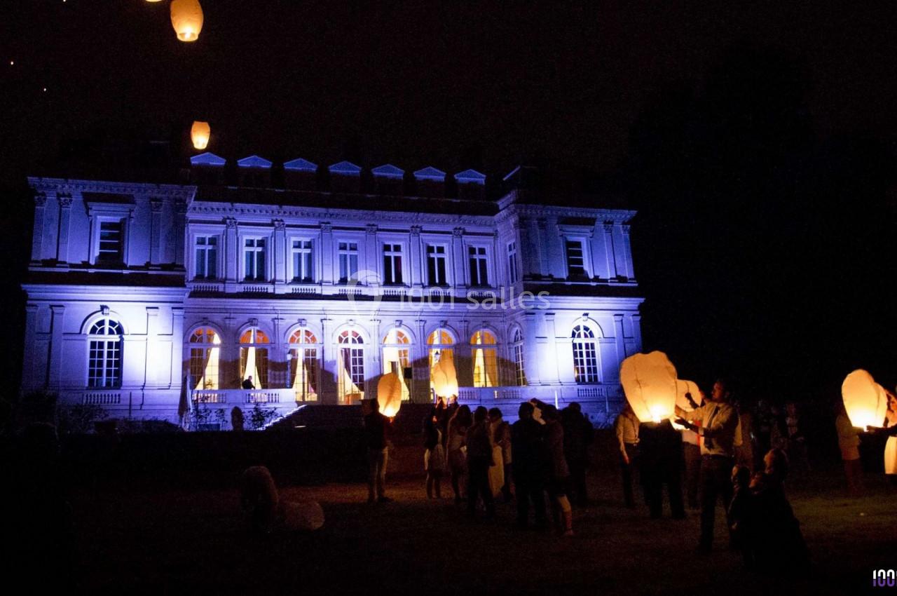 Des personnes lâchent des lanternes lumineuses devant un grand bâtiment éclairé en bleu, de nuit.
