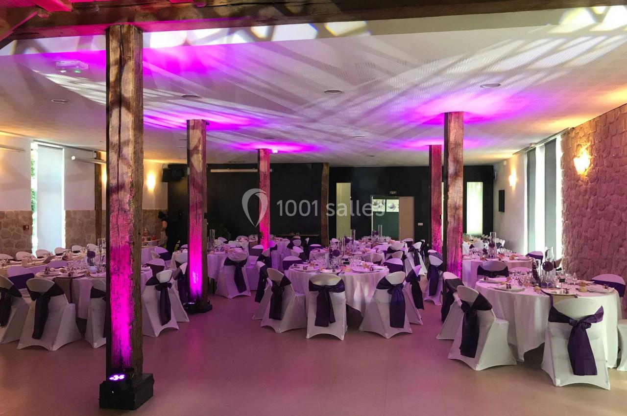 Salle de réception décorée avec des tables rondes dressées, nappes blanches et chaises ornées de rubans violets.
