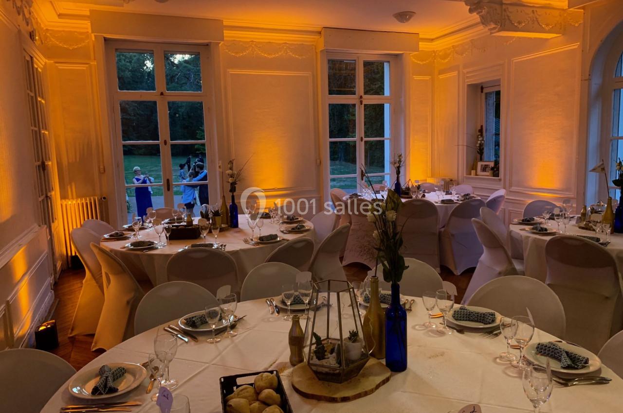 Salle élégamment décorée pour un dîner, avec tables rondes dressées, éclairage chaleureux et vue sur un jardin.