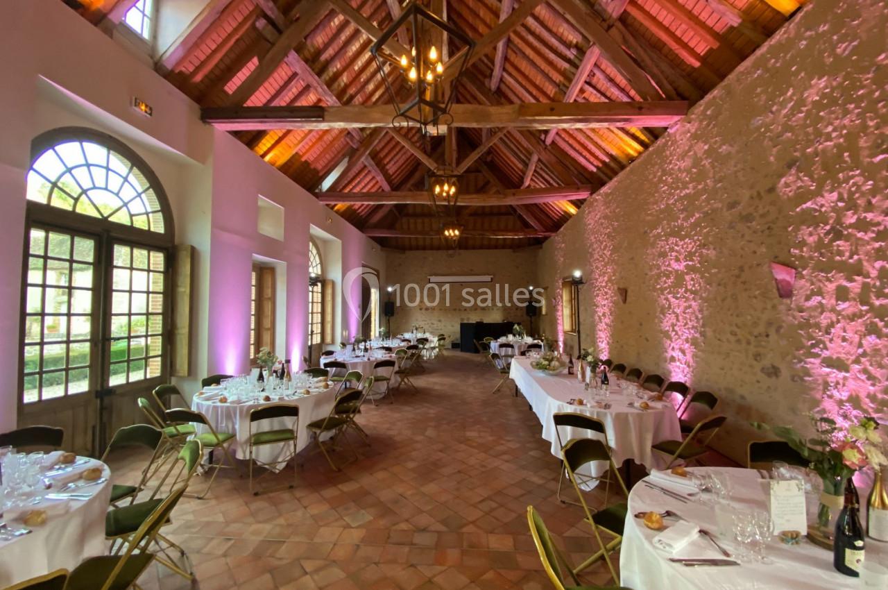 Salle de réception avec tables rondes dressées, chaises pliantes et éclairage chaleureux sous une charpente en bois.