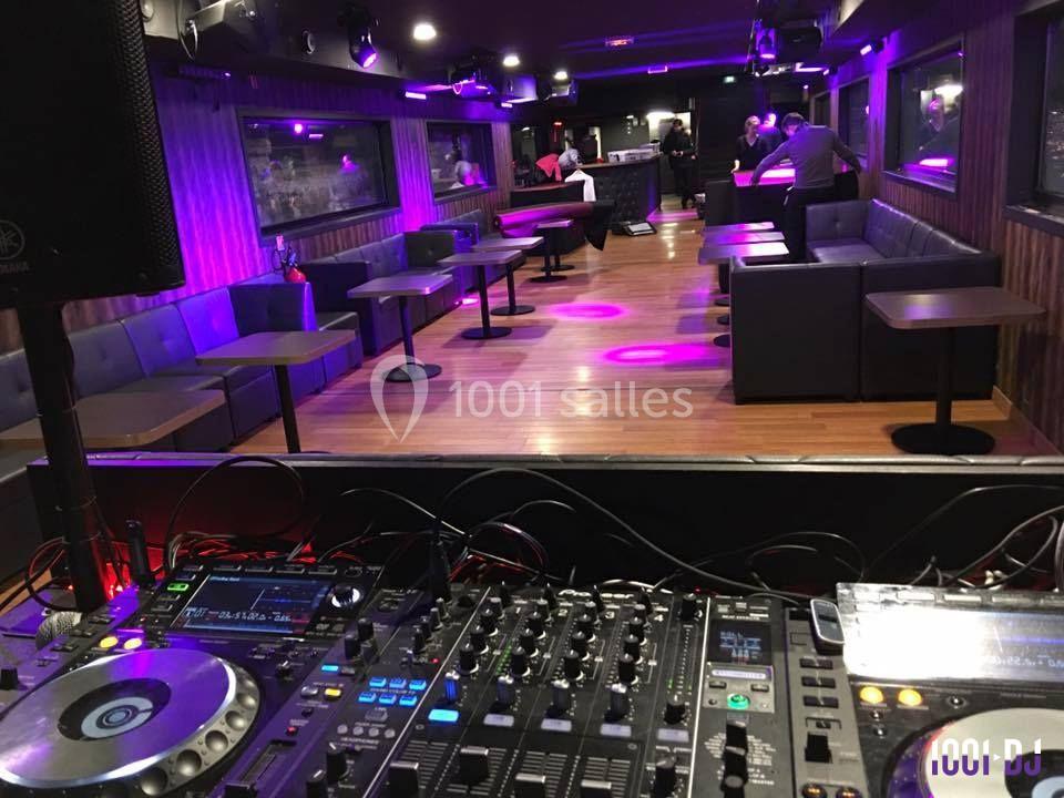 Vue d'une salle de réception avec tables et banquettes, éclairée par des lumières violettes, depuis une console de DJ.
