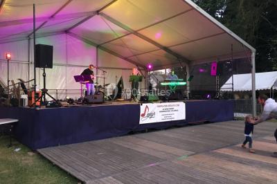 Groupe de musiciens jouant sur une scène couverte en plein air, éclairée par des lumières colorées, avec un public devant.