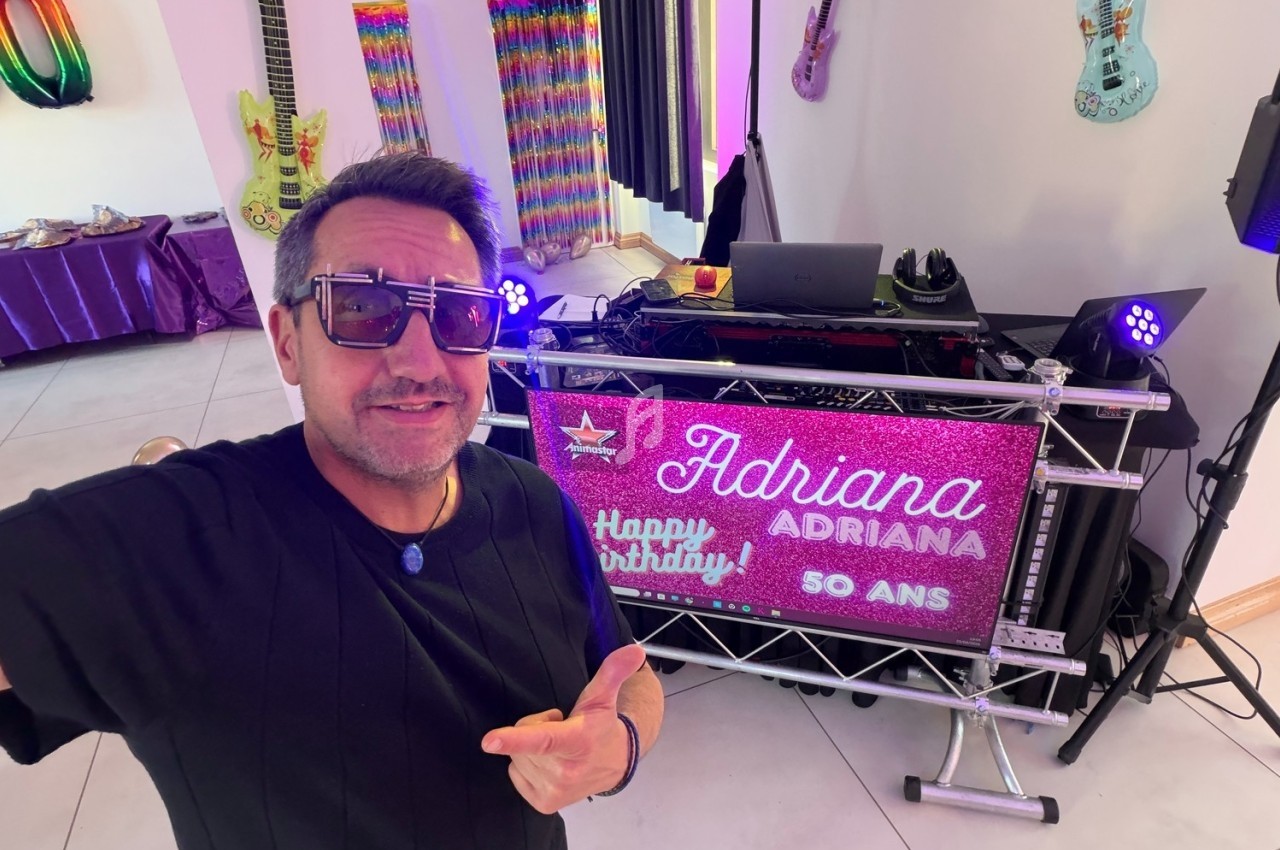 Homme souriant prenant un selfie devant une table de DJ décorée pour les 50 ans d'Adriana, avec des guitares au mur.