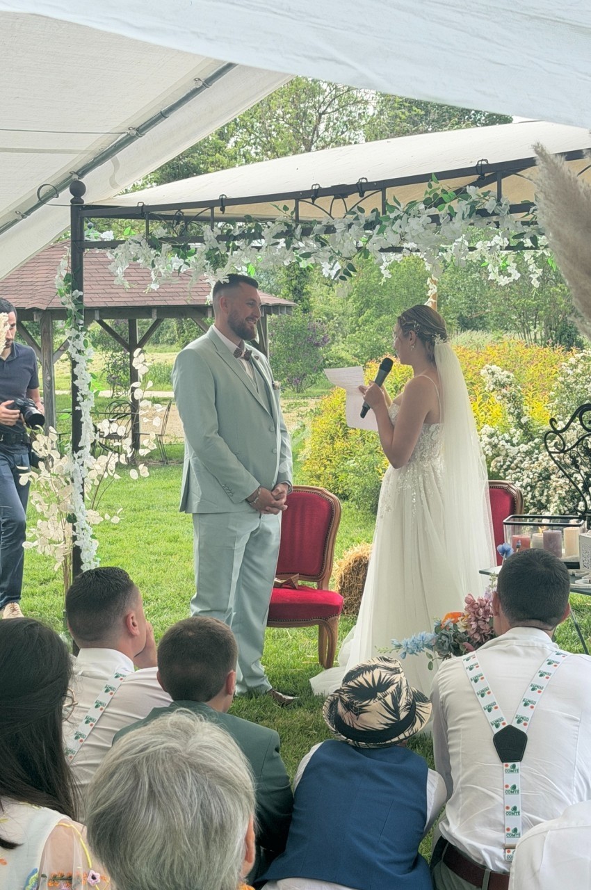 Une mariée en robe blanche s'adresse à son époux debout sous une tente lors d'une cérémonie en extérieur.