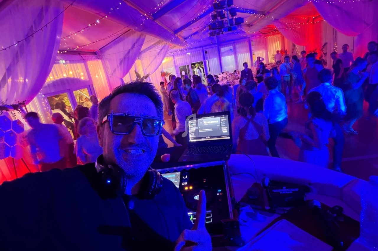 Un DJ souriant anime une soirée dansante dans une salle éclairée par des lumières colorées, avec des invités sur la piste.