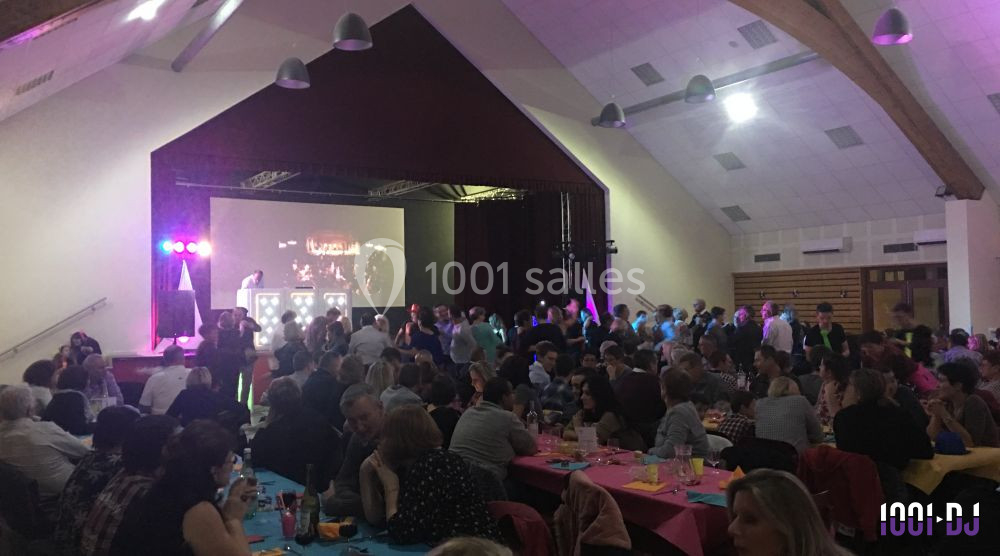 Salle comble avec des tables décorées, des convives assis et une scène éclairée avec un écran et des animations.