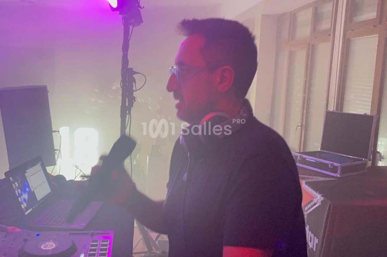 Un DJ tenant un micro devant une table de mixage, éclairé par des lumières colorées dans une ambiance enfumée.