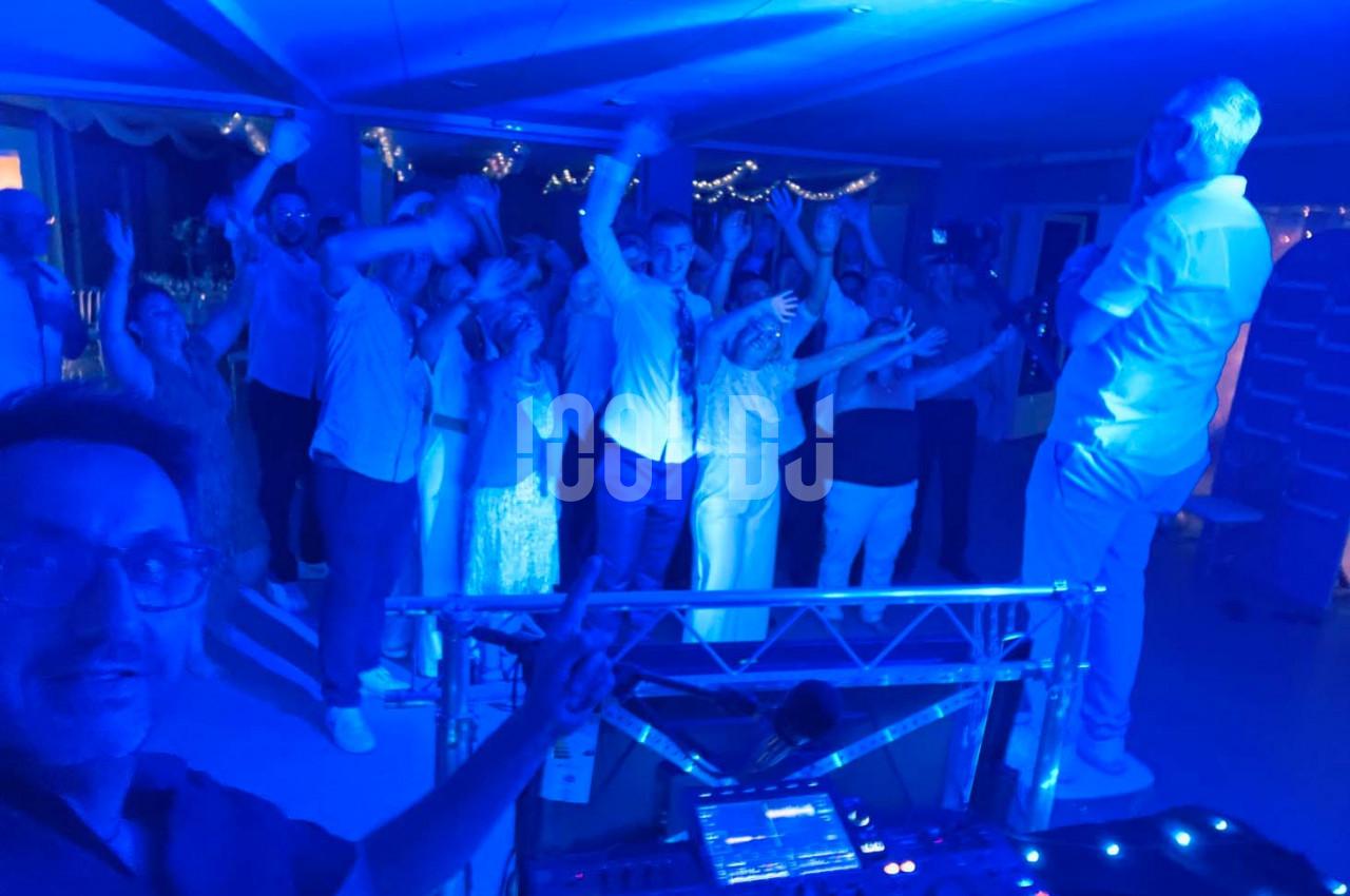 Groupe de personnes dansant sous des lumières bleues lors d'une fête, avec un DJ au premier plan.