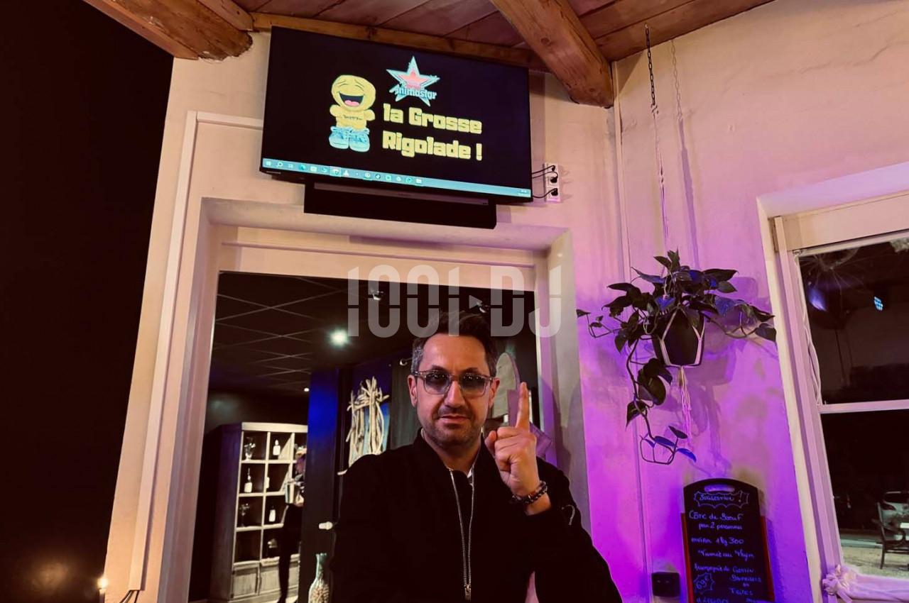 Un homme pose devant un écran affichant ’La Grosse Rigolade !’ dans un intérieur avec plantes et ardoise décorative.