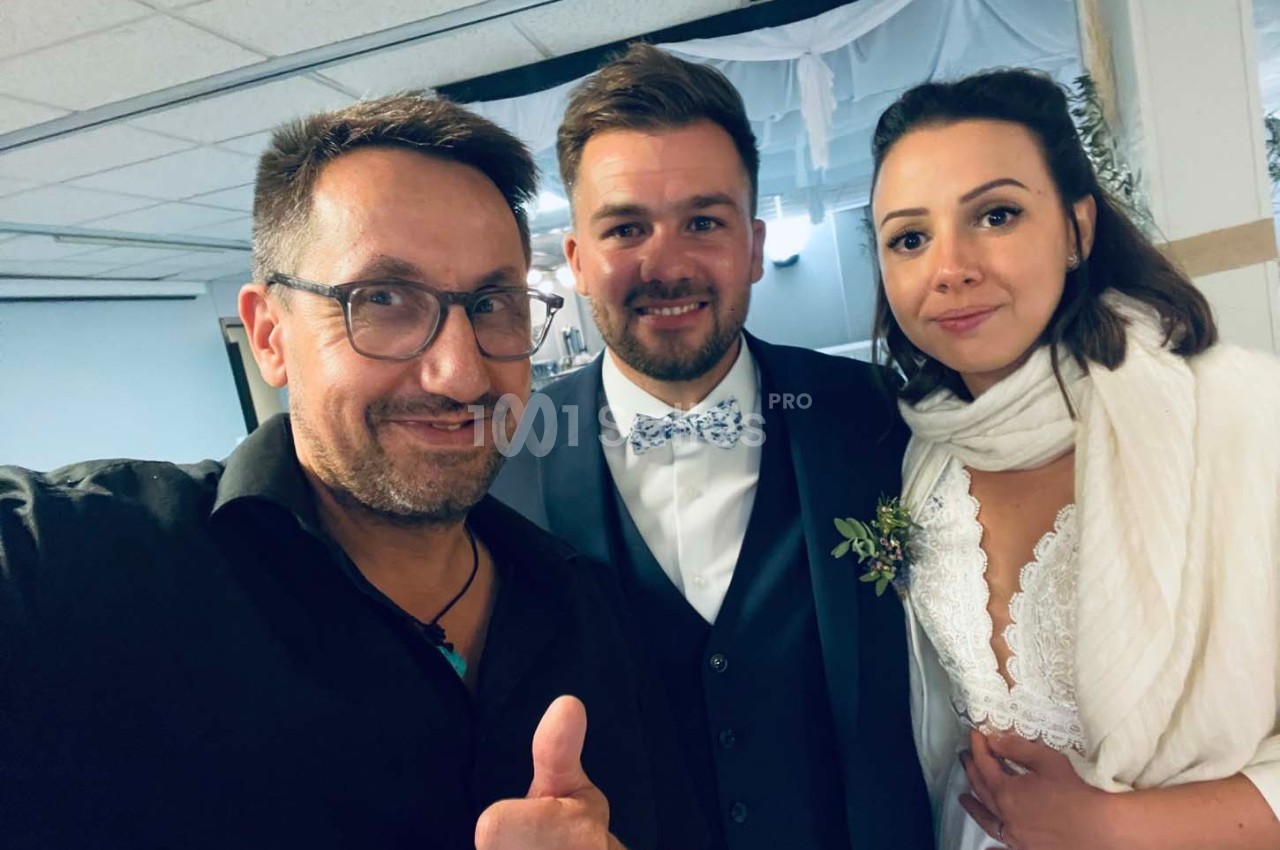 Un homme souriant prend un selfie avec un couple habillé en tenue de mariage dans une salle décorée.