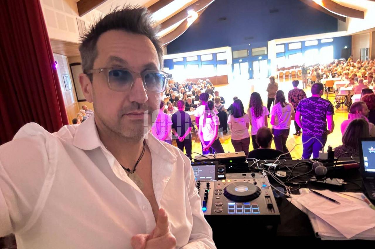 Un homme en chemise blanche devant une table de mixage, face à un public dans une salle lumineuse.