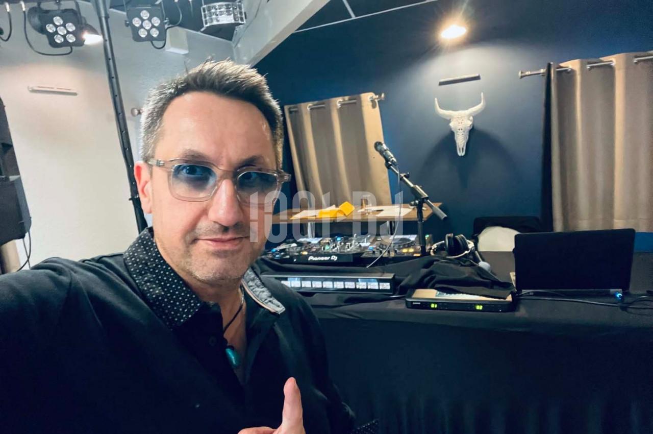 Homme posant devant une table de mixage avec des équipements audio, un micro et une décoration murale en arrière-plan.