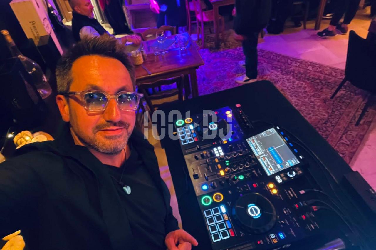 Un homme pose devant une table de mixage DJ dans un environnement intérieur avec éclairage tamisé.