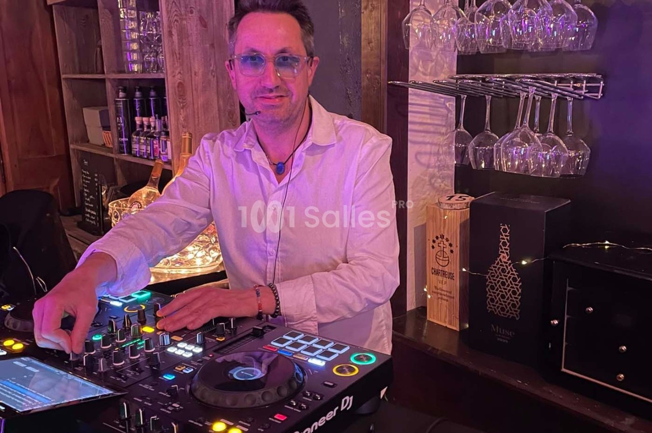 Un DJ en chemise blanche mixe de la musique sur une console dans un bar avec des verres et des bouteilles en arrière-plan.