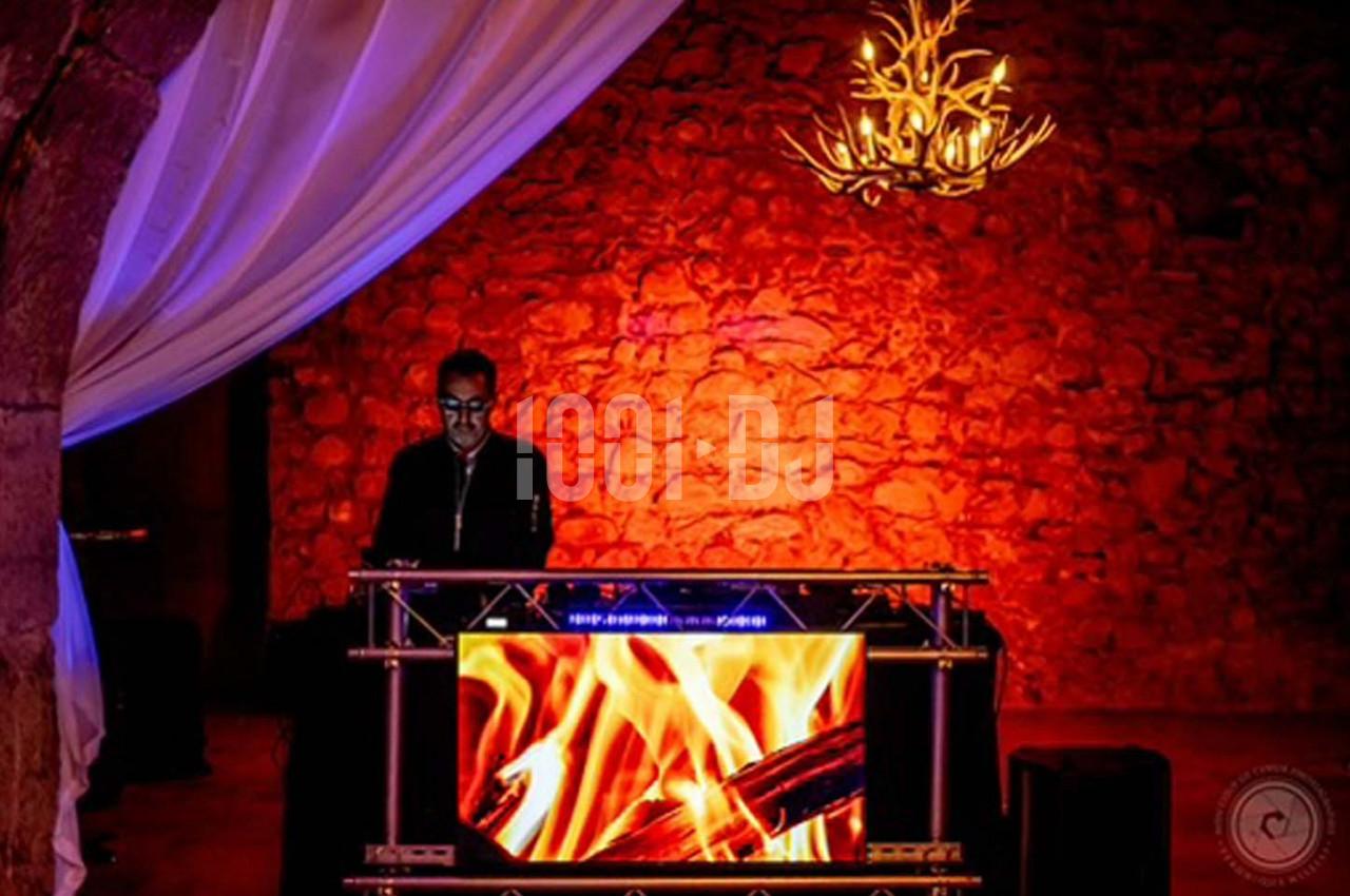 Un DJ mixe derrière une table ornée d'un écran affichant des flammes, dans une salle aux murs en pierre éclairée en rouge.