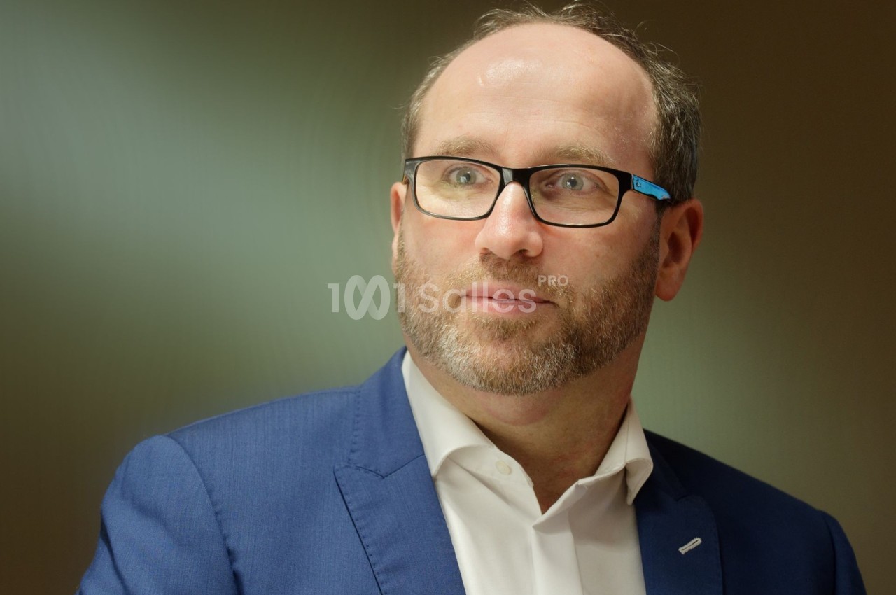 Homme portant des lunettes et un costume bleu, regardant vers la droite dans un environnement intérieur.