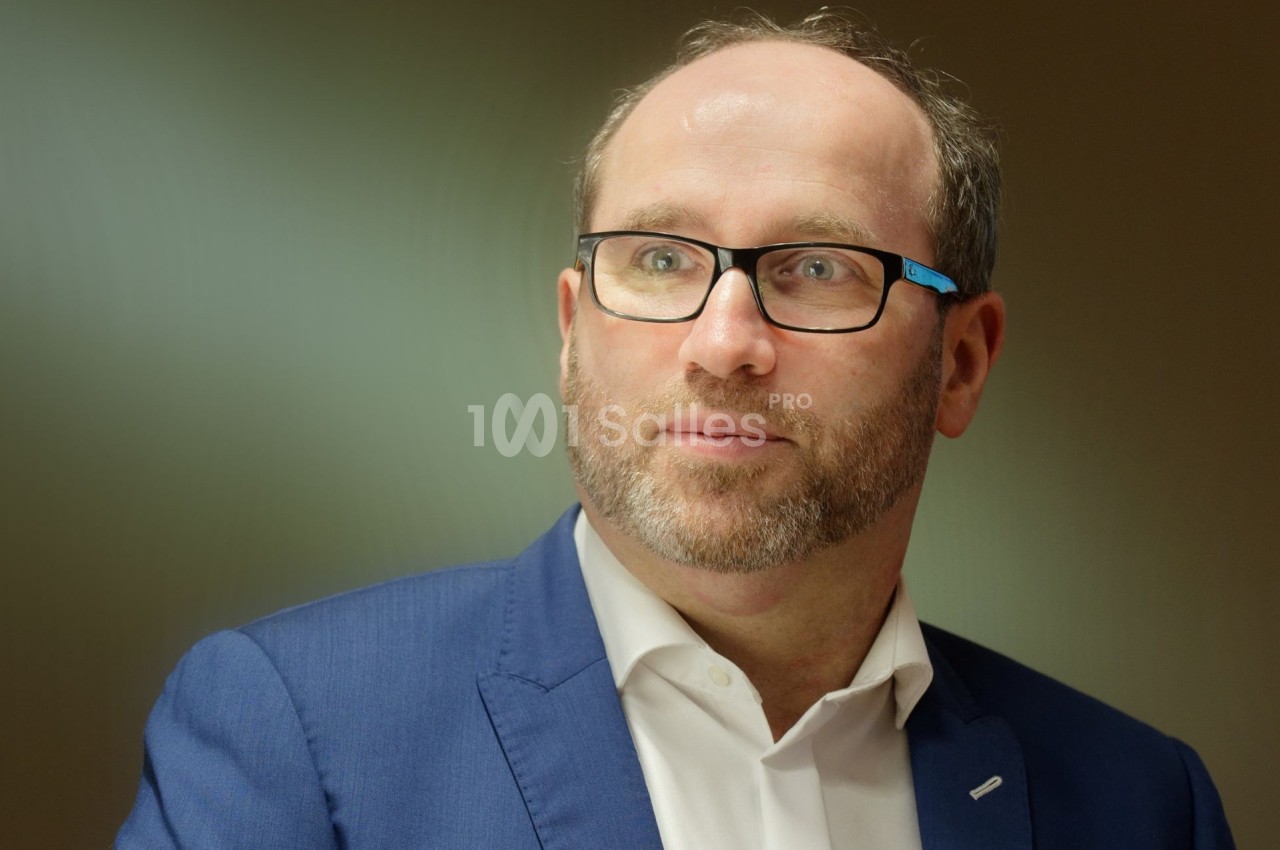 Homme portant des lunettes et un costume bleu, regardant légèrement sur le côté dans un environnement intérieur.