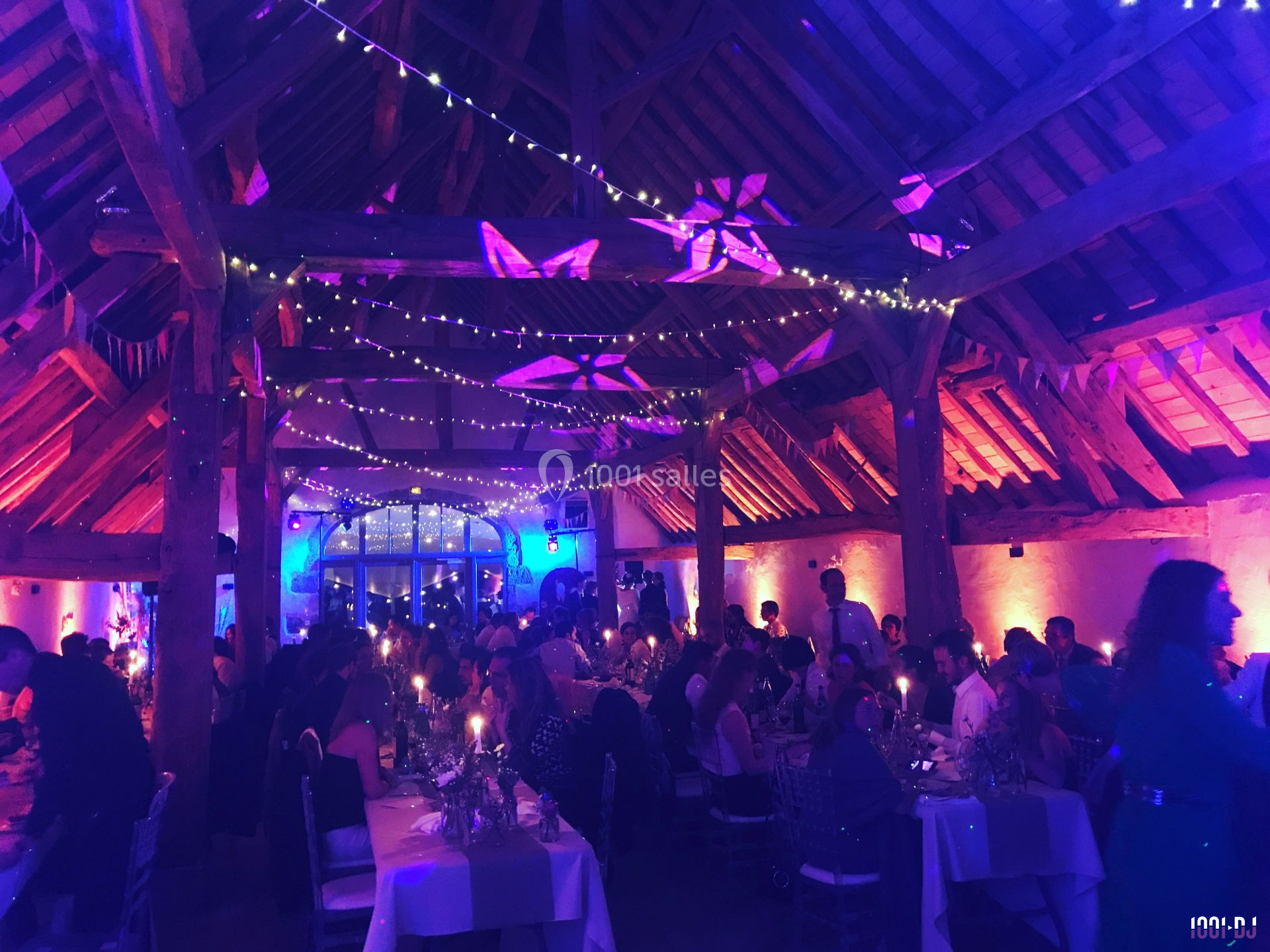 Salle de réception décorée avec des guirlandes lumineuses, remplie d'invités assis à des tables lors d'un événement.