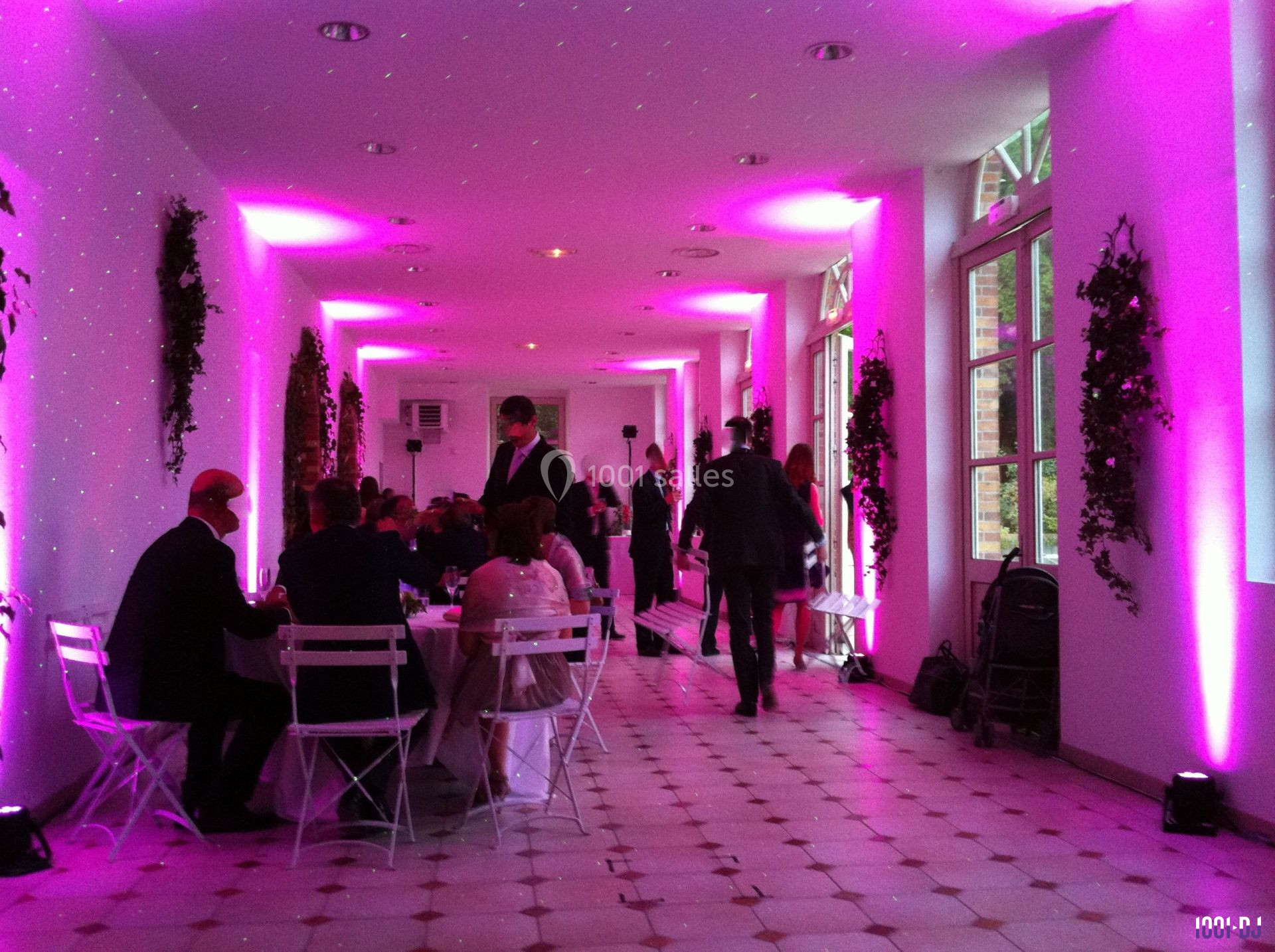 Salle éclairée de lumières roses, avec des invités assis à des tables blanches et d'autres debout dans un couloir.