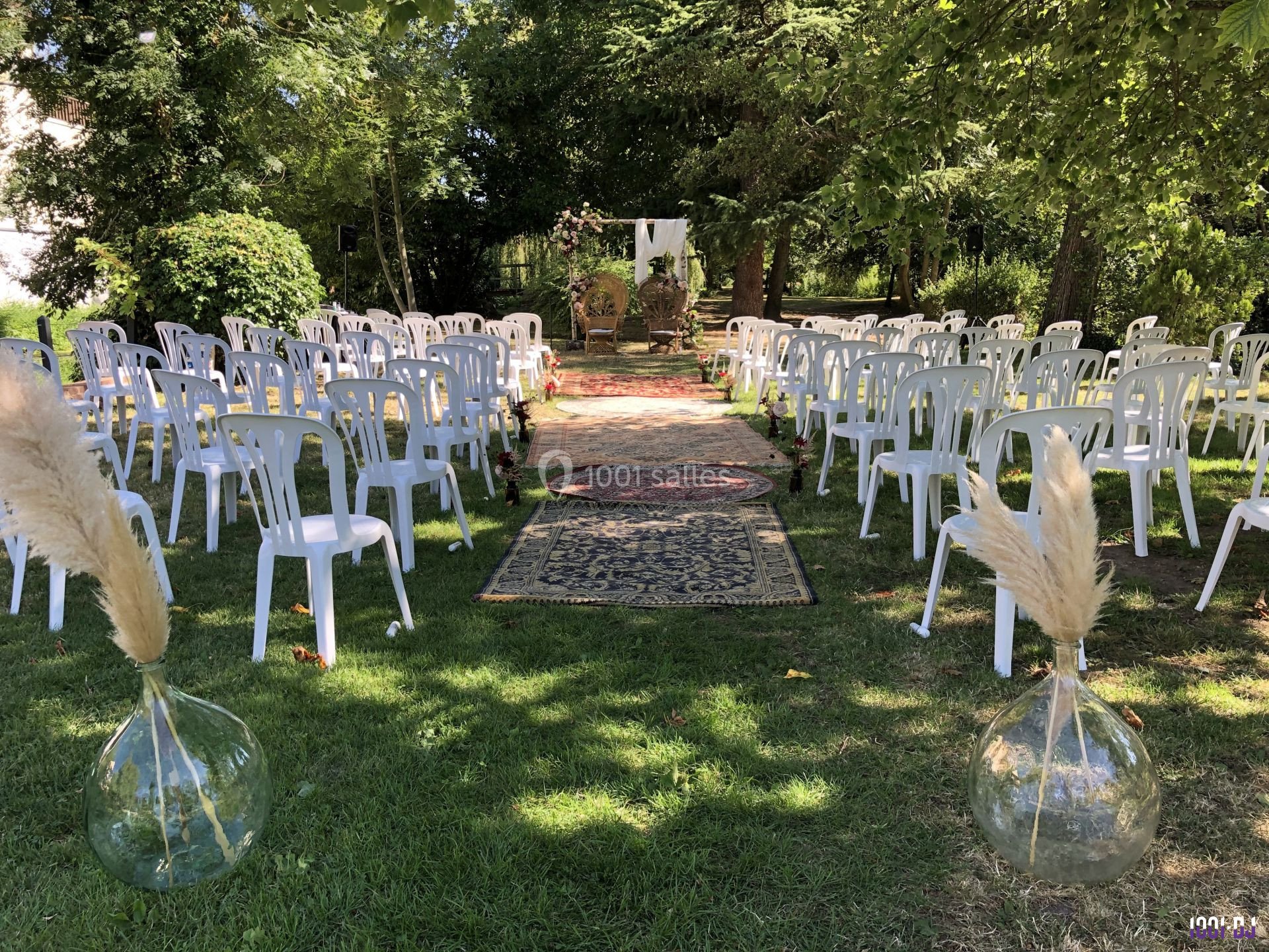 Chaises blanches disposées en extérieur, formant une allée centrale décorée de tapis et de vases avec des herbes séchées.