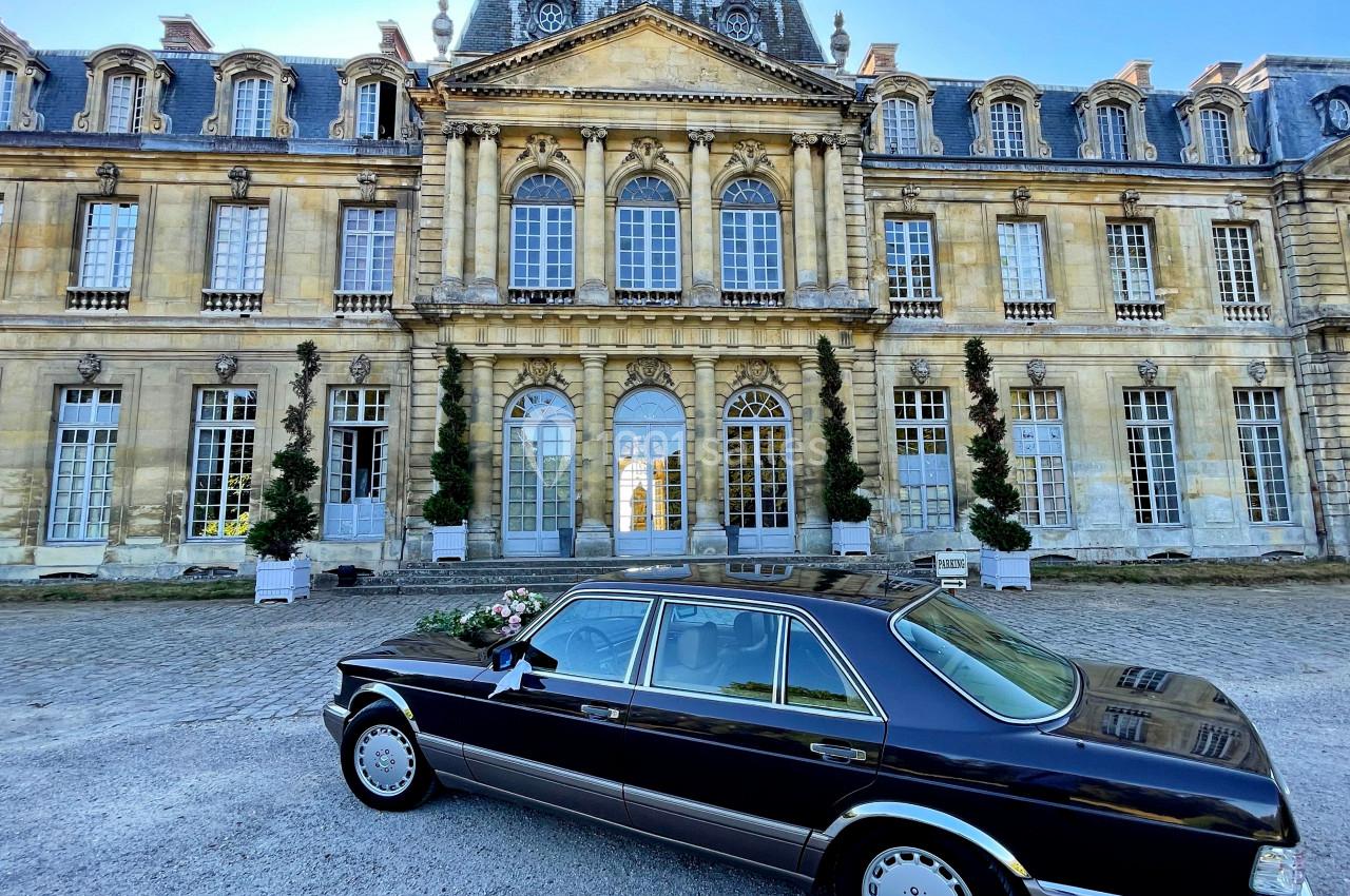 Voiture noire garée devant un château en pierre avec des fenêtres à arcades et un toit mansardé.