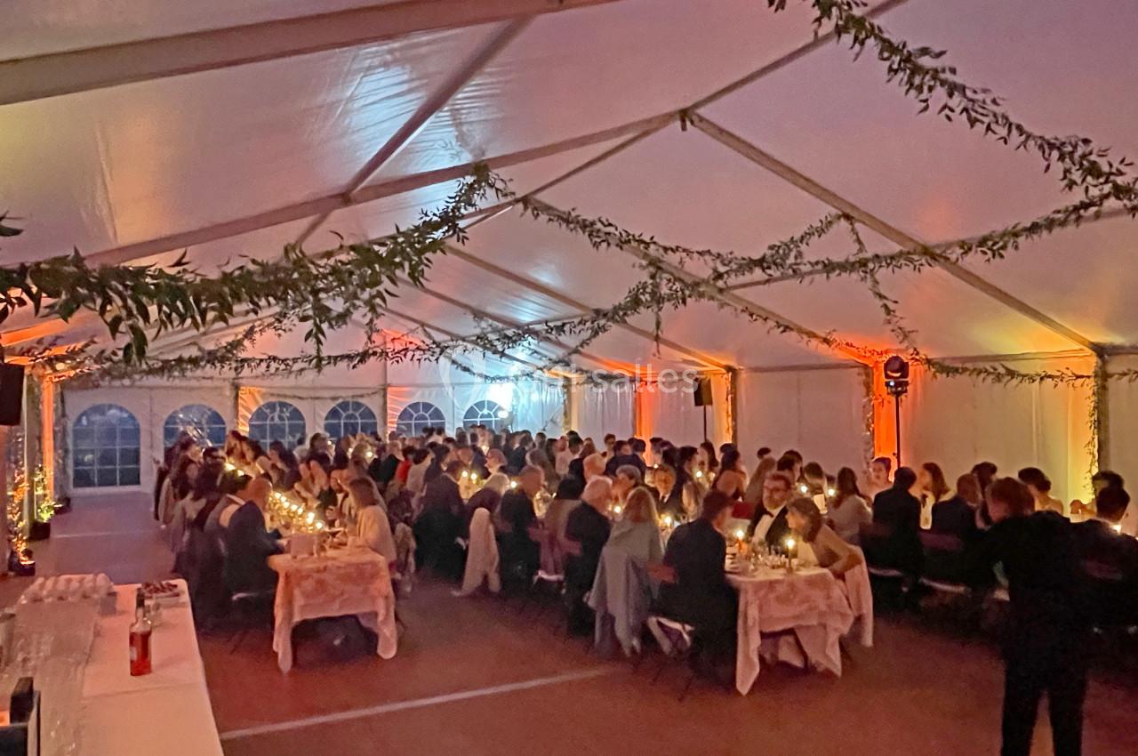 Dîner festif sous une grande tente éclairée, avec des convives assis à des tables décorées et illuminées.