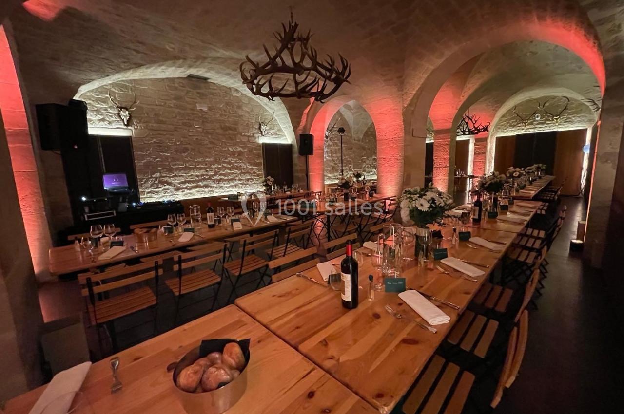 Salle voûtée éclairée en tons chauds, aménagée avec de longues tables en bois décorées pour un dîner.