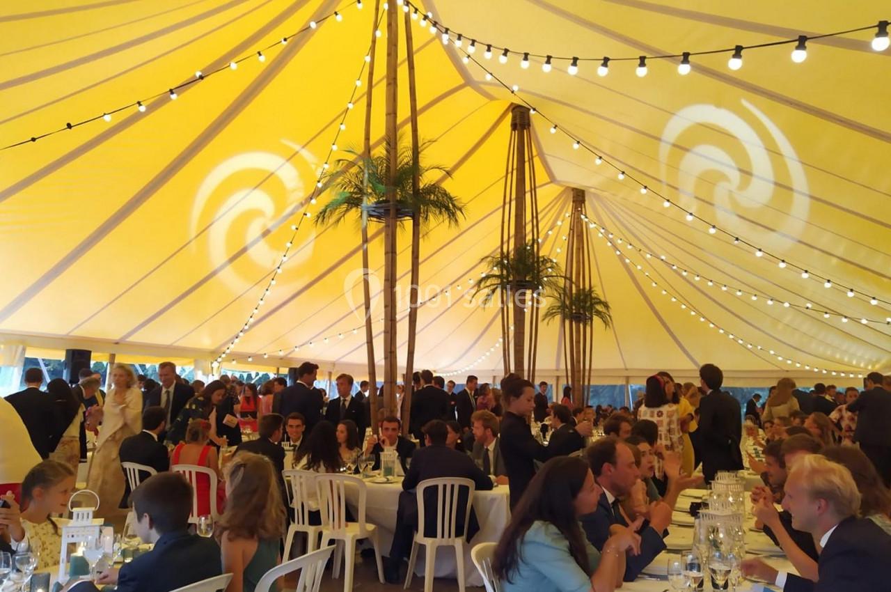 Invités assis et debout sous une grande tente éclairée, décorée de guirlandes lumineuses et de faux palmiers.