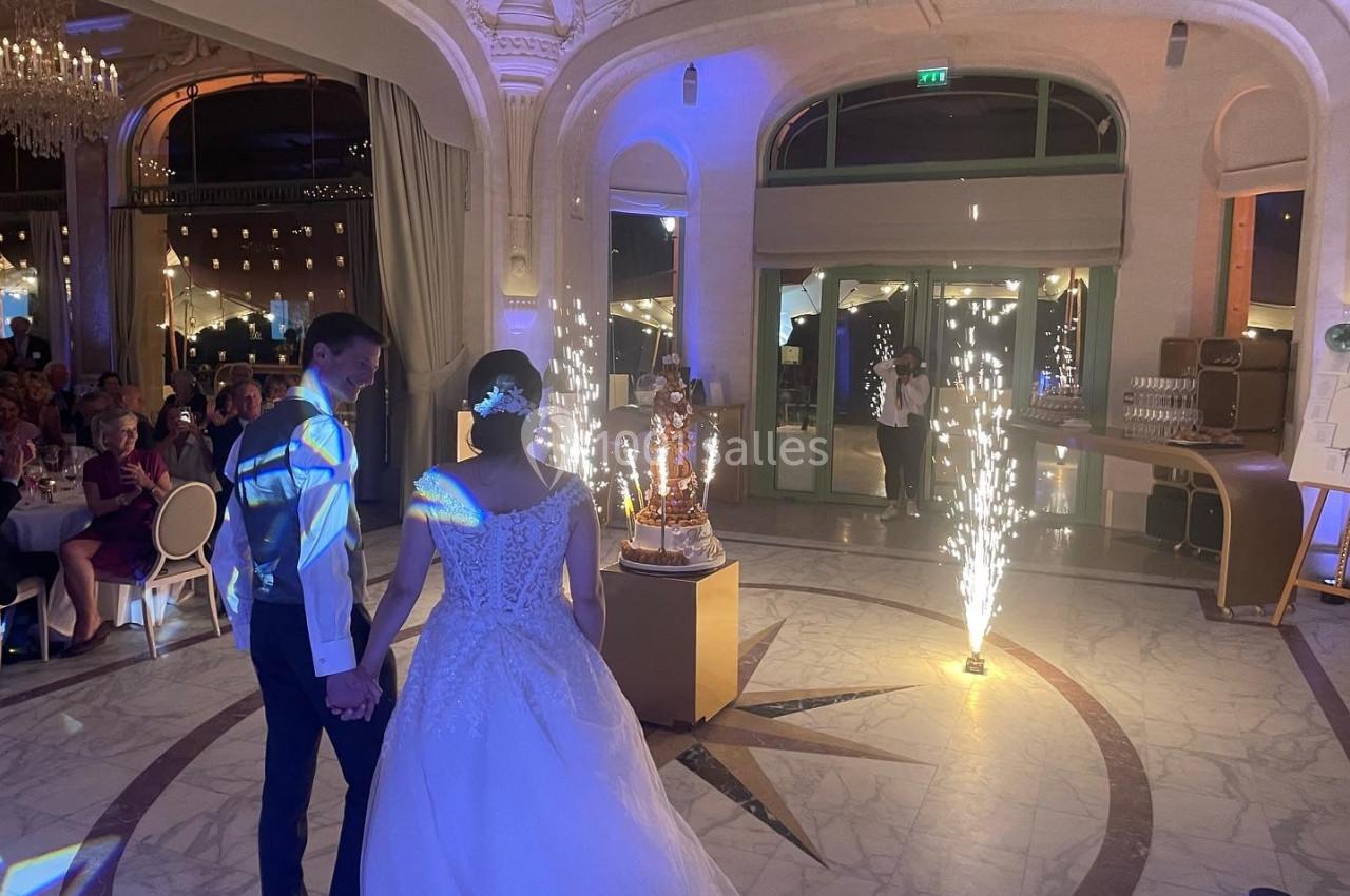Un couple en tenue de mariage regarde une pièce montée illuminée par des étincelles dans une salle élégante.