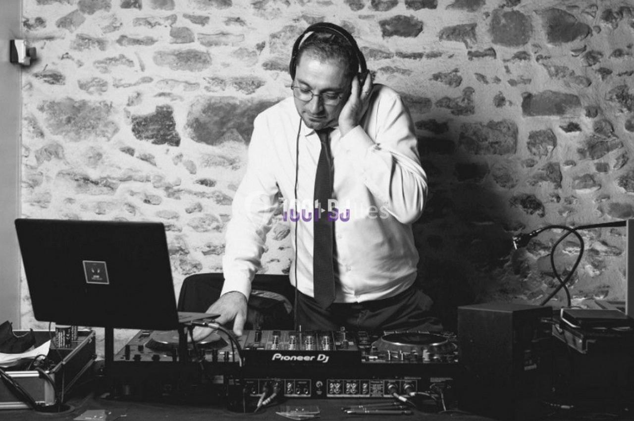 Un DJ en chemise et cravate mixe de la musique sur une table de mixage devant un mur en pierre.