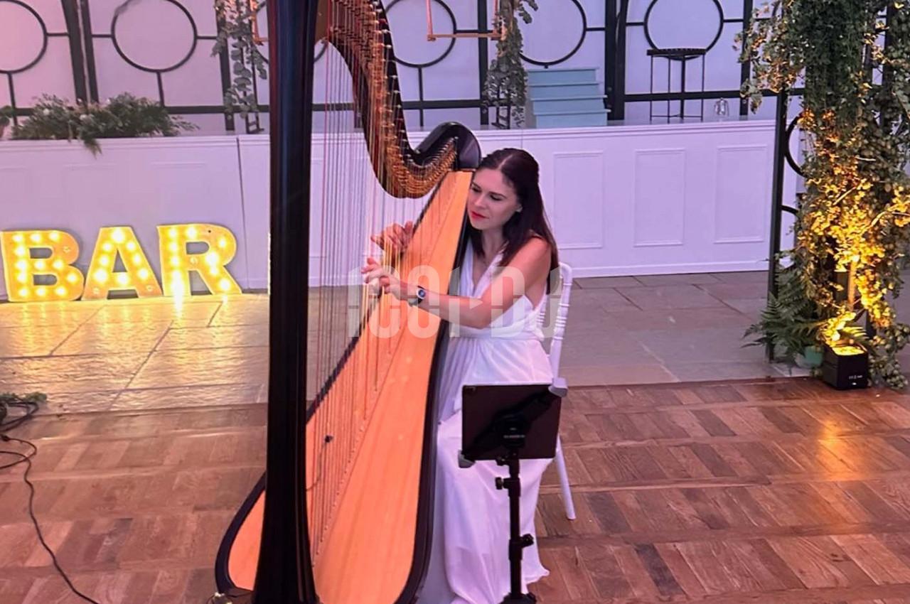 Une femme en robe blanche joue de la harpe dans une salle décorée avec des plantes et des lumières.
