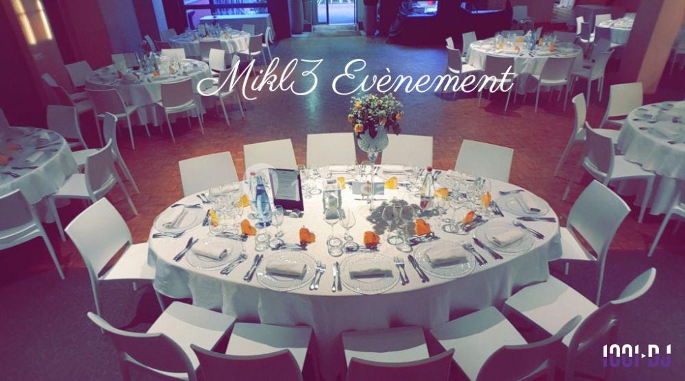 Salle décorée pour un événement avec des tables rondes dressées, nappes blanches et un centre de table floral.