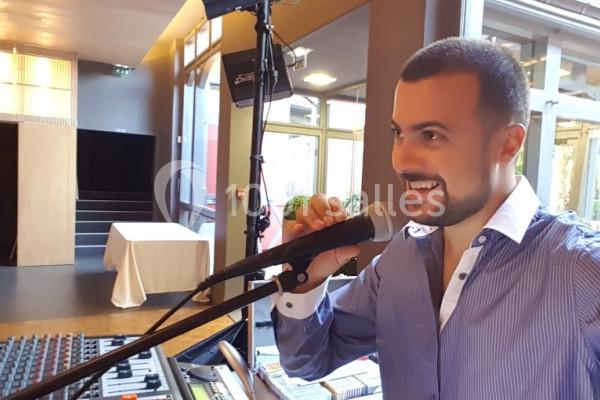 Homme souriant parlant dans un micro, devant une table de mixage dans un espace intérieur lumineux.