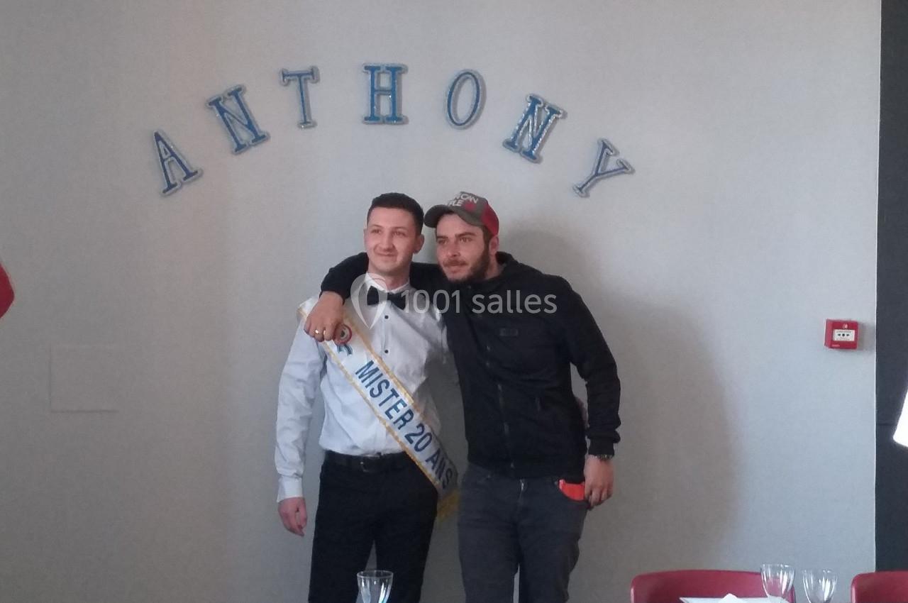 Deux hommes posent côte à côte dans une salle, l'un portant une écharpe ’Mister 2015’ et un nœud papillon.