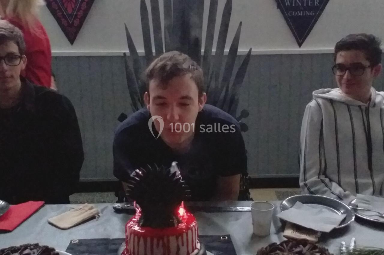 Un jeune homme souffle une bougie sur un gâteau décoré, entouré de deux personnes assises à une table.