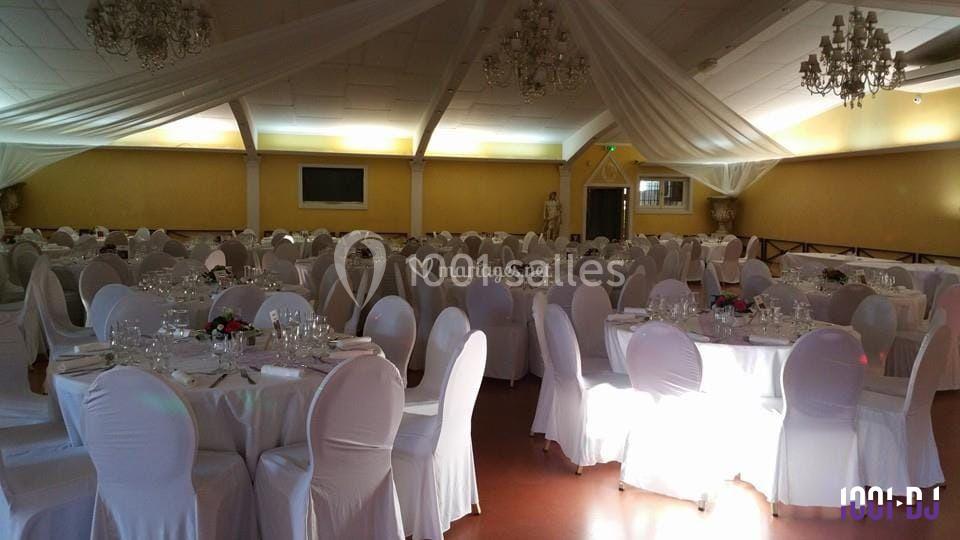 Salle de réception décorée avec des tables rondes, nappes blanches, chaises houssées et lustres suspendus.