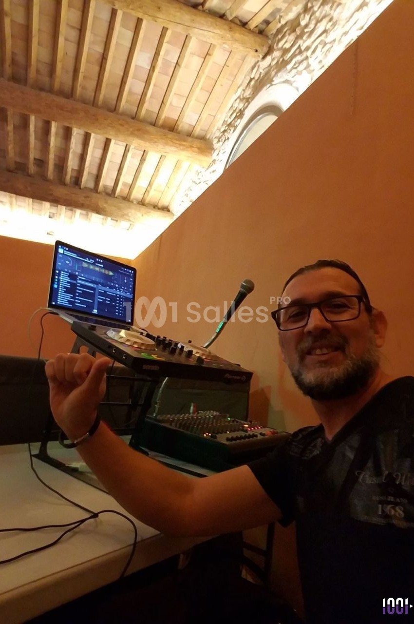 Homme souriant devant un équipement de DJ avec ordinateur, console de mixage et microphone dans une pièce au plafond en bois.