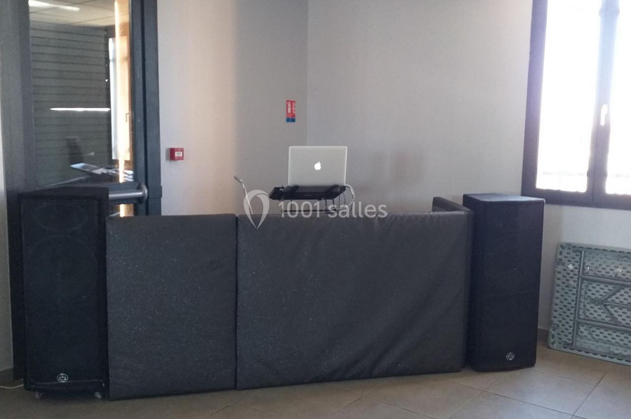 Poste de DJ avec ordinateur portable, enceintes et table recouverte de panneaux noirs dans une pièce lumineuse.