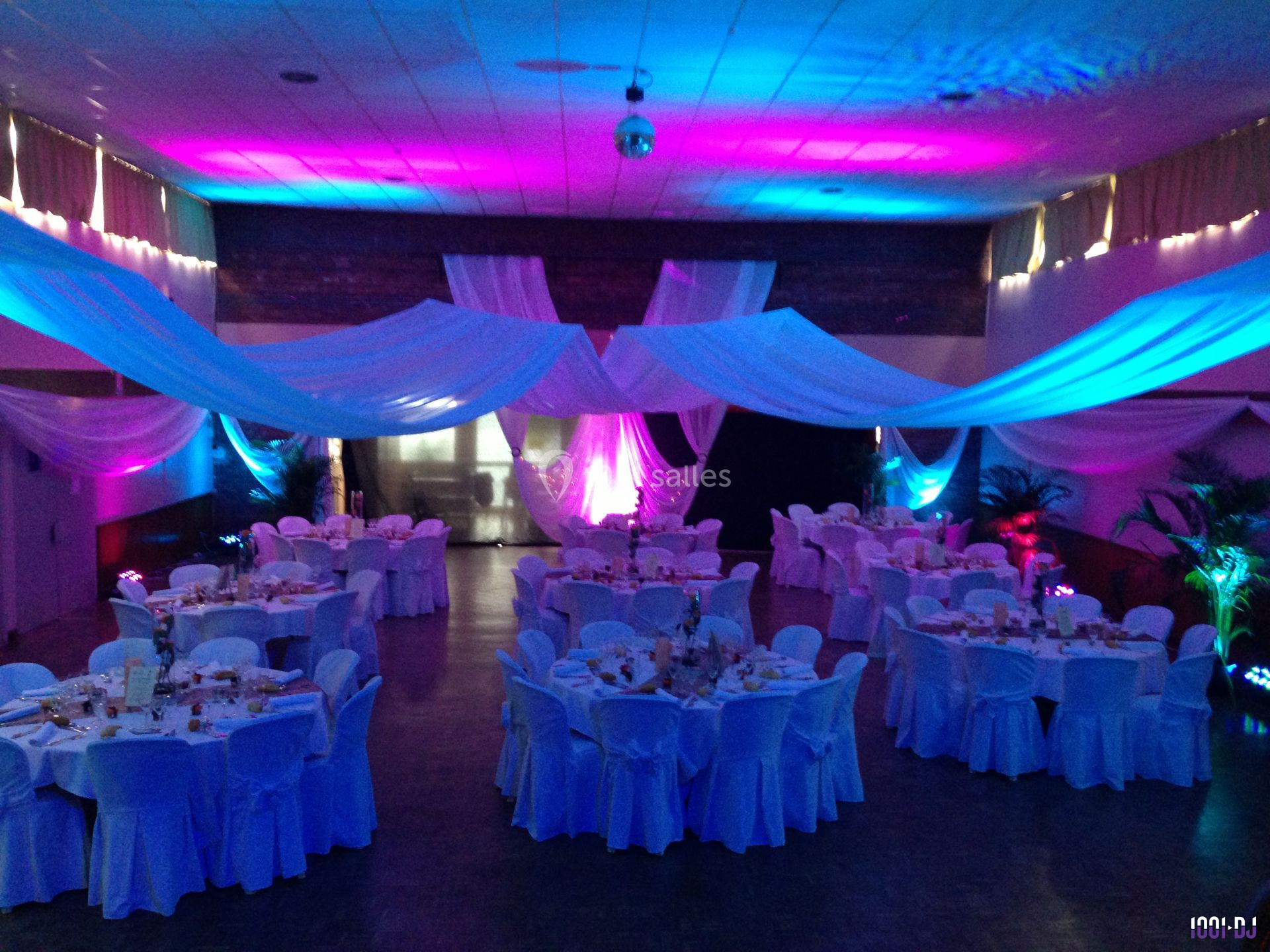 Salle de réception décorée avec des tables rondes, nappes blanches et éclairages colorés créant une ambiance festive.