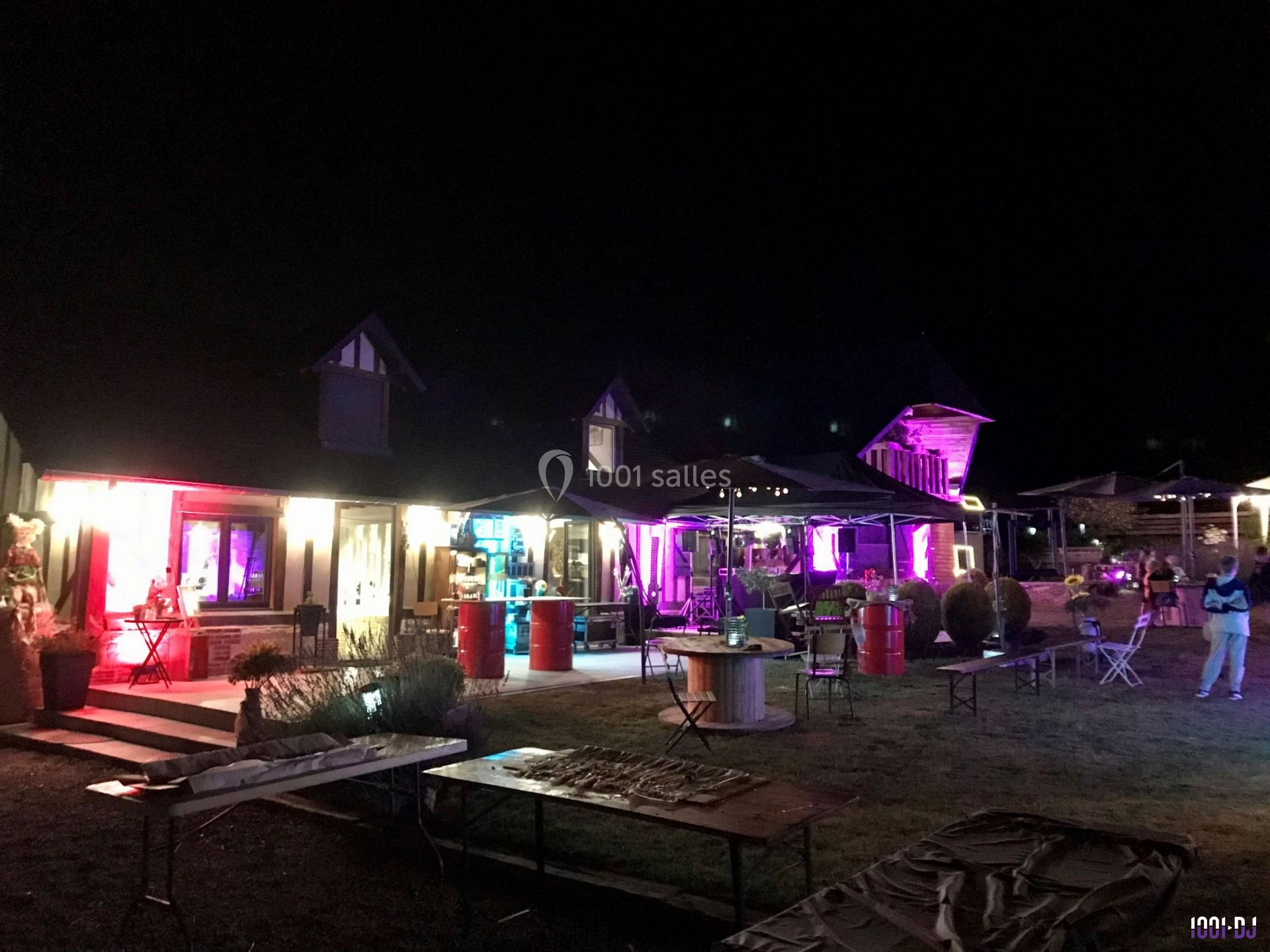 Bâtiment éclairé par des lumières colorées la nuit, avec des tables et parasols disposés à l'extérieur.