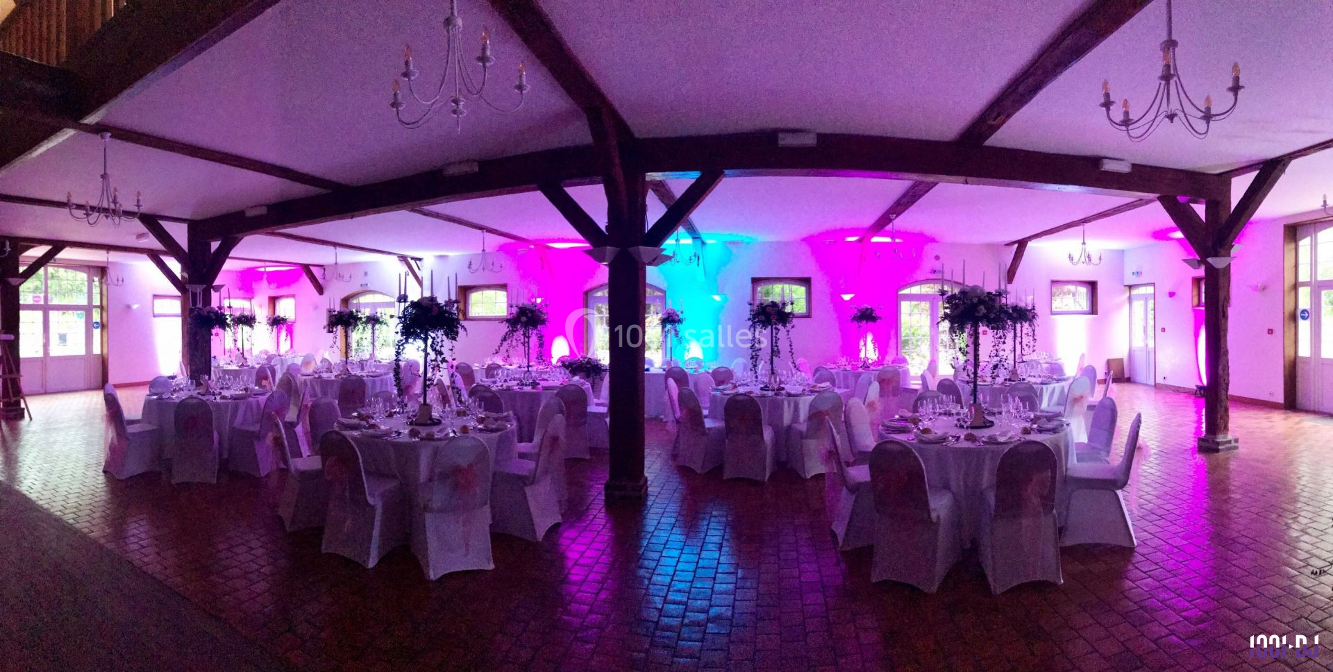 Salle de réception décorée avec des tables rondes, nappes blanches, centres de table fleuris et éclairage coloré.