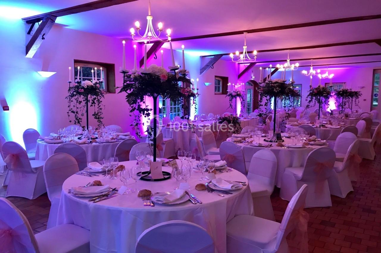 Salle de réception décorée avec des tables rondes, nappes blanches, chaises ornées de rubans roses et éclairage tamisé.