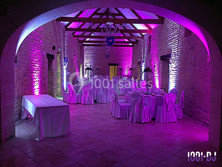 Salle de réception en pierre éclairée de lumières violettes, avec tables rondes dressées et chaises décorées.