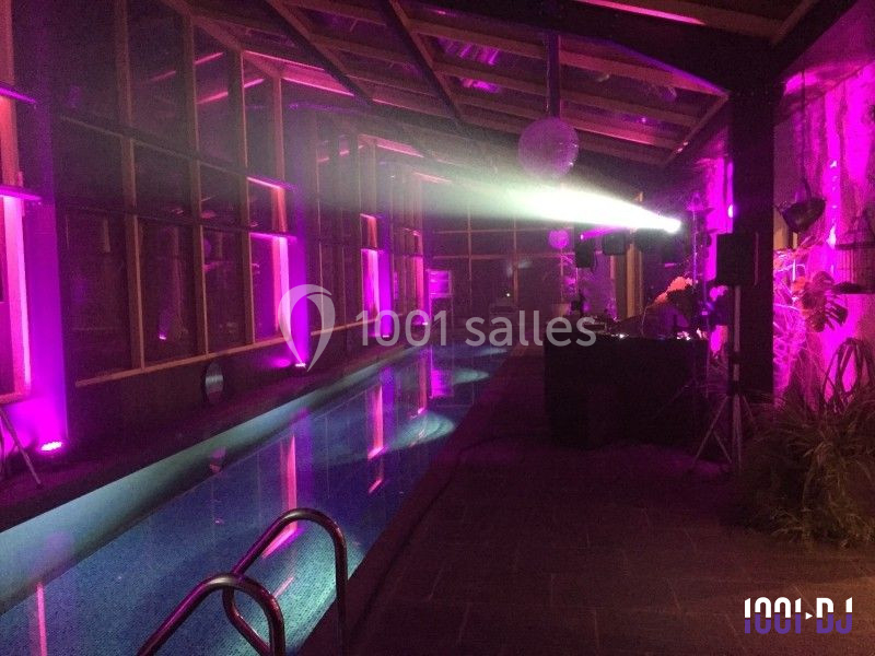 Salle avec piscine éclairée par des lumières violettes, DJ et équipement audio installés sur le côté.