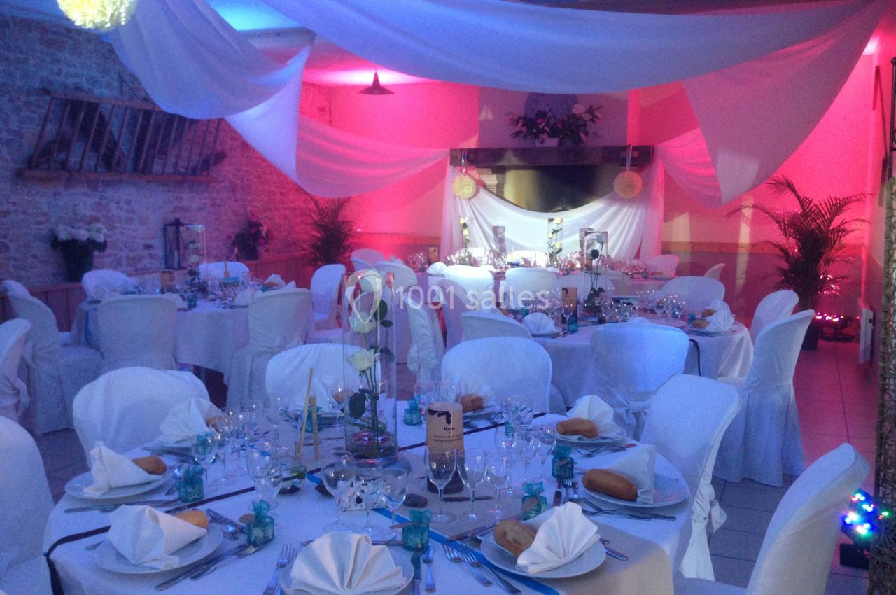 Salle de réception décorée avec des nappes blanches, des tables dressées et un éclairage rose et bleu.