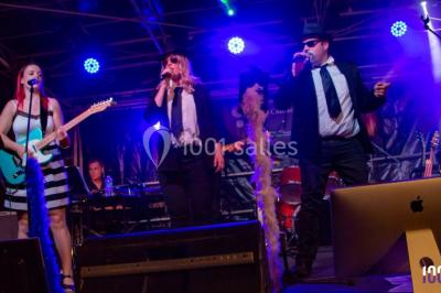 Scène éclairée avec une table de mixage, des enceintes et des affiches en arrière-plan dans une salle intérieure.